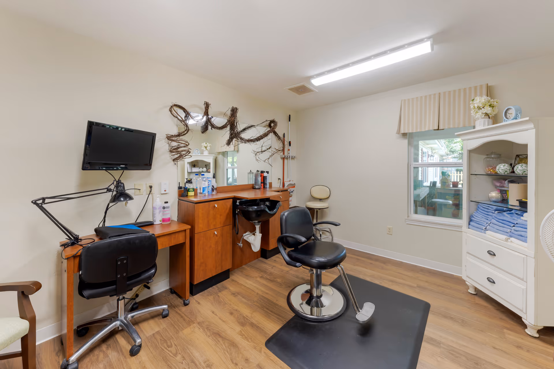 Interior salon room with styling chairs, a wash basin, mirror, TV and cabinets on wood flooring.