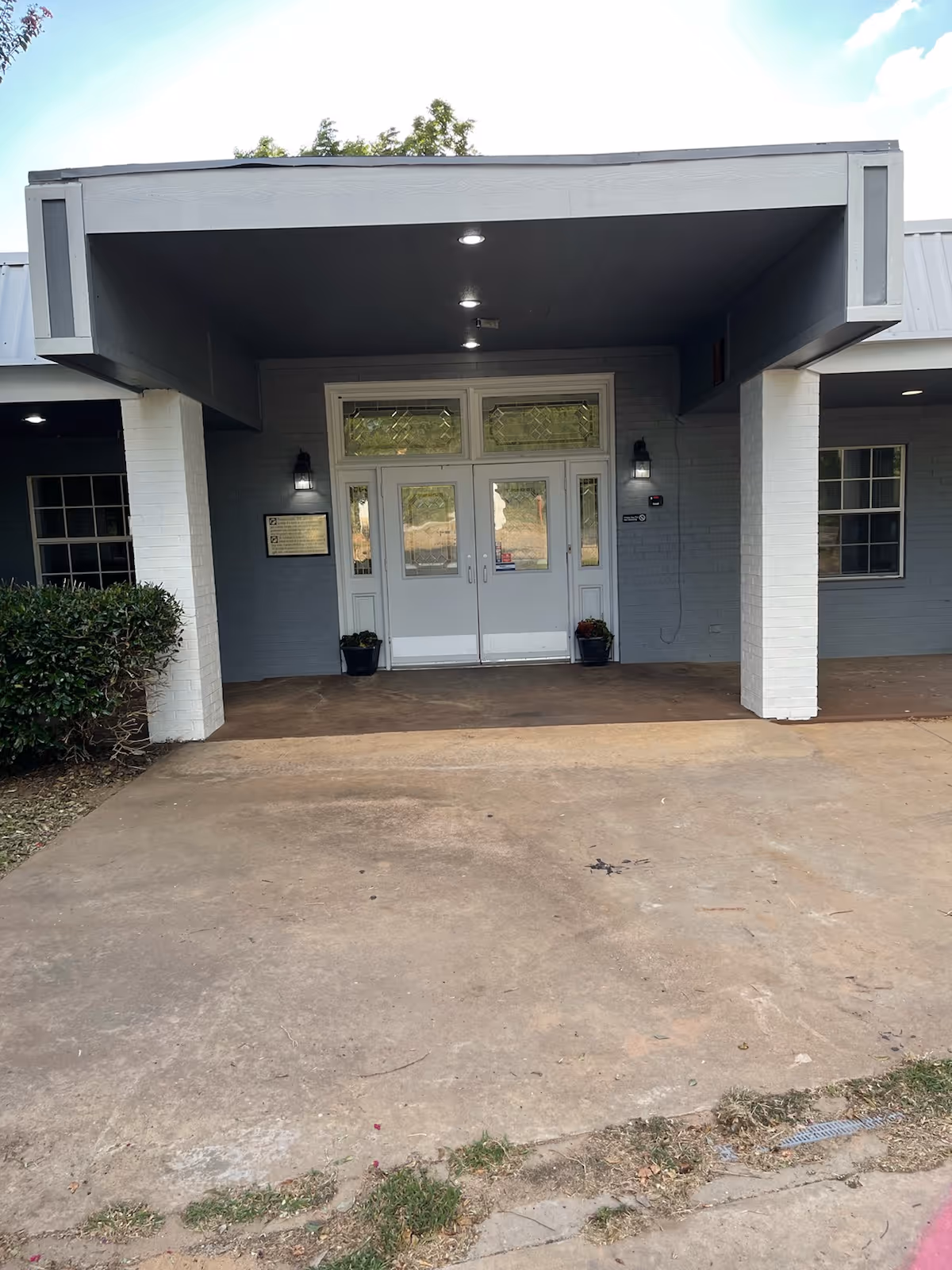 Entrance of a building with a covered porch area supported by white pillars. The building has double glass doors with decorative glass panels above and on the sides. There are two wall-mounted lights on either side of the doors and two potted plants placed near the entrance. The ground is paved with concrete and there is some greenery visible on the left side.
