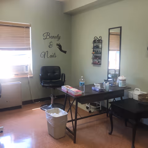 A small beauty and nail care room with a black salon chair, a table holding various supplies including tissues, a water bottle, and nail polish remover. A wall-mounted rack with nail polish bottles is next to a tall mirror. The wall has a decal that reads 'Beauty & Nails'. A window with blinds and an air conditioning unit is visible on the left side.