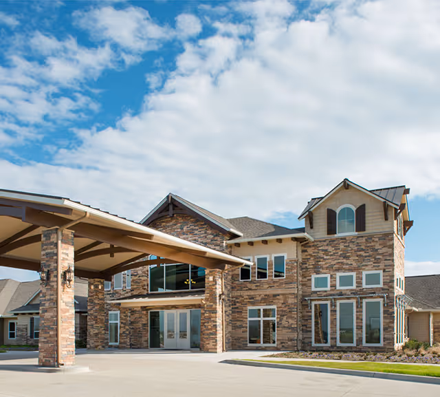 Exterior view of a large assisted living facility building with stone facade, multiple windows, and a covered entrance under a partly cloudy blue sky.