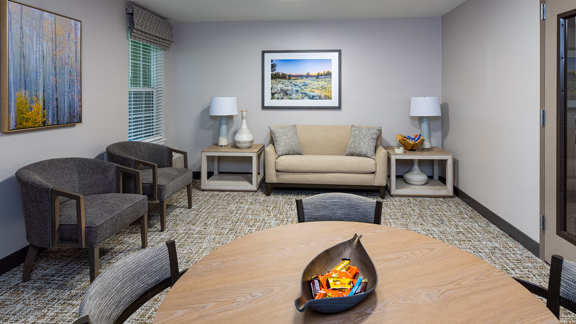 A cozy sitting area in a senior living facility with a beige sofa flanked by two side tables each holding a lamp and decorative items. Two gray armchairs are positioned to the left near a window with blinds and a valance. A round wooden table with chairs is in the foreground, featuring a decorative bowl filled with candy. The walls are light gray with a framed landscape photograph above the sofa and a colorful abstract painting on the left wall. The floor is carpeted with a patterned design.