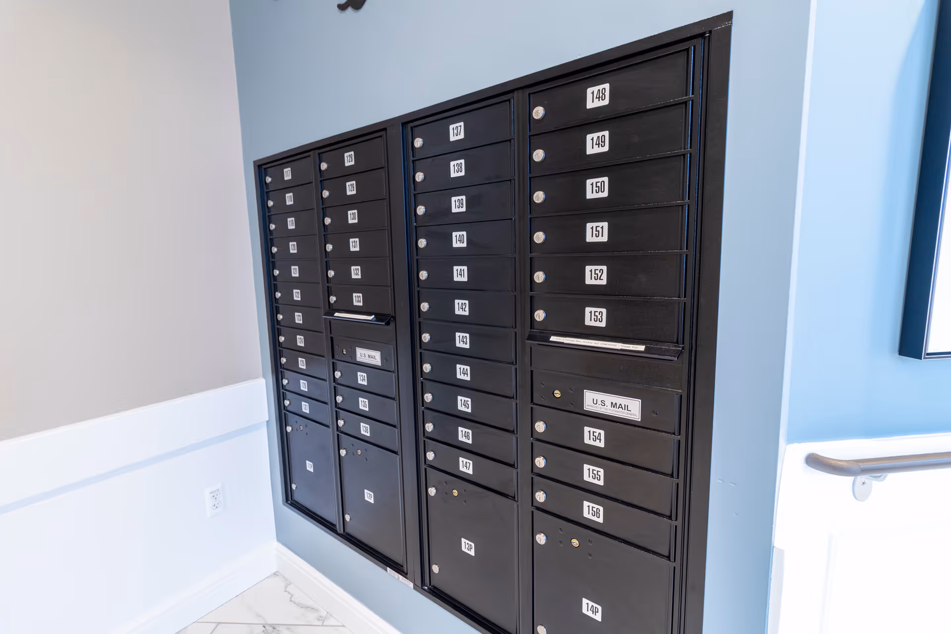 A wall-mounted bank of black U.S. mailboxes with numbered slots in a light-blue interior hallway.