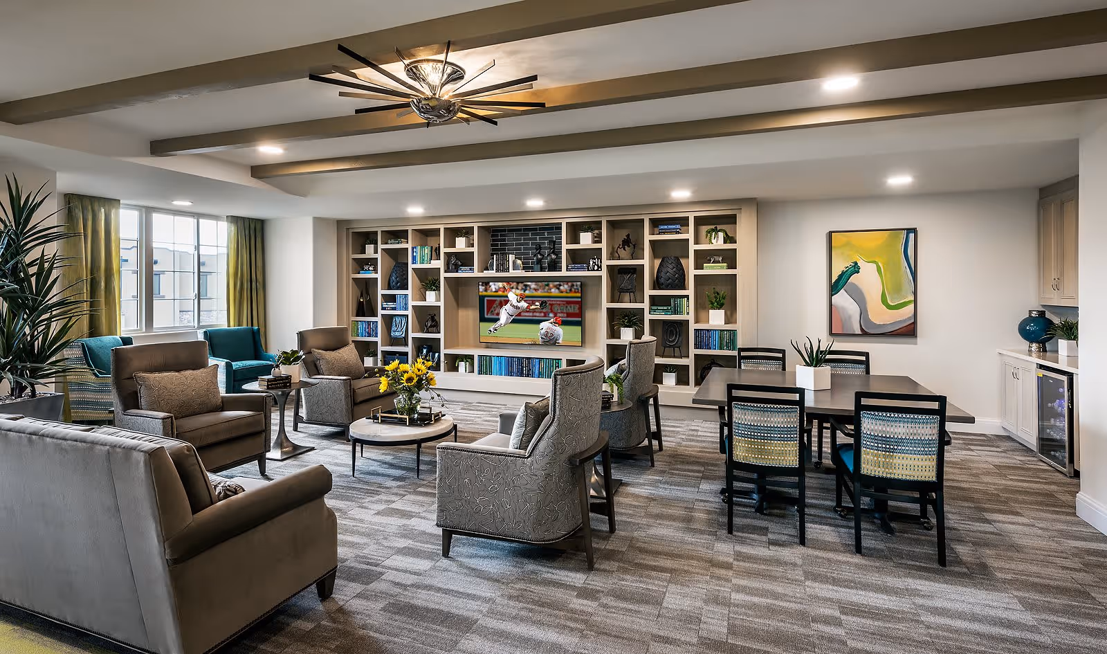 A spacious and well-lit living room area with multiple armchairs and a sofa arranged around a round coffee table with a vase of yellow flowers. There is a large built-in shelving unit with decorative items and a TV showing a baseball game. A dining table with six chairs is positioned near a colorful abstract painting on the wall. The room has large windows with green curtains, a modern ceiling fan, and a carpeted floor.