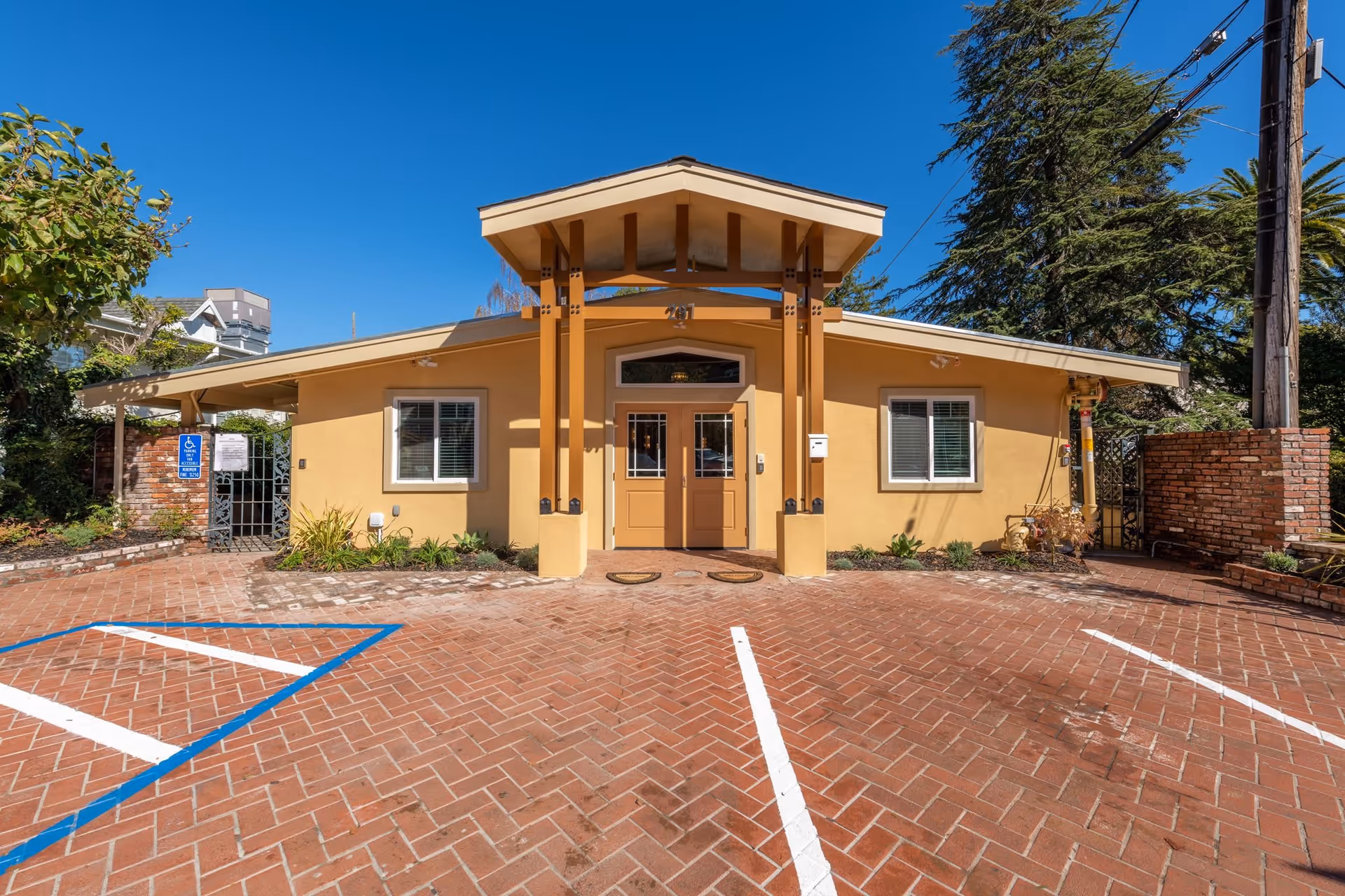 Front entrance of a single-story yellow stucco building with double doors, decorative columns and a brick parking area with marked spaces.