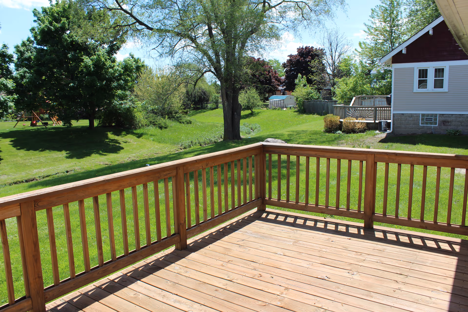 Wooden deck with railing overlooking a grassy backyard, trees, and a neighboring house.
