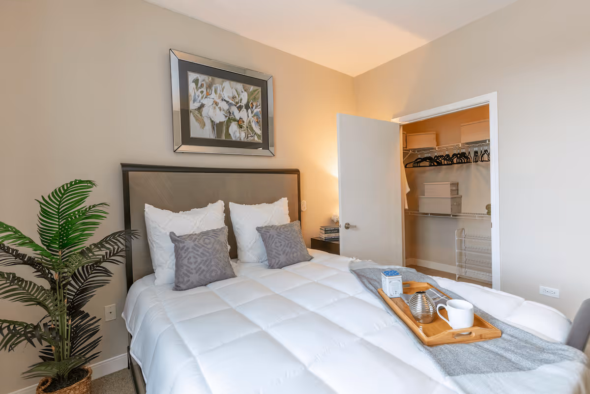A neatly made bed with white bedding and gray pillows in a bedroom. A wooden tray with a teapot, cup, and other items is placed on the bed. There is a potted plant to the left of the bed and a framed floral painting on the wall above the headboard. An open door reveals a closet with hangers and storage boxes inside.