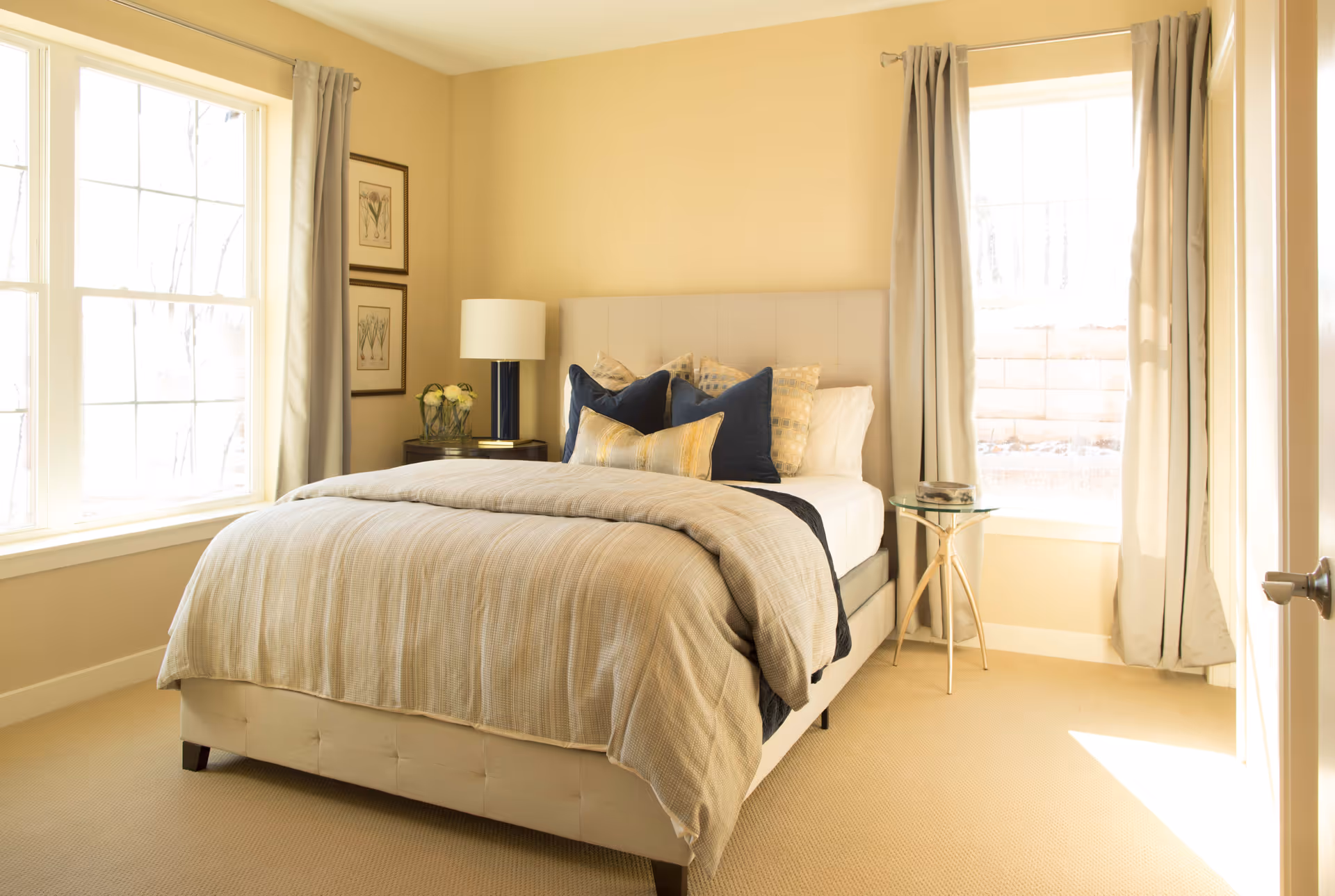 A bright and cozy bedroom with a neatly made bed featuring beige and navy blue pillows and a beige comforter. The room has two large windows with light curtains, a small round glass side table on the right, and a dark wooden nightstand with a lamp and flower vase on the left. The walls are painted a soft yellow, and the carpet is beige.