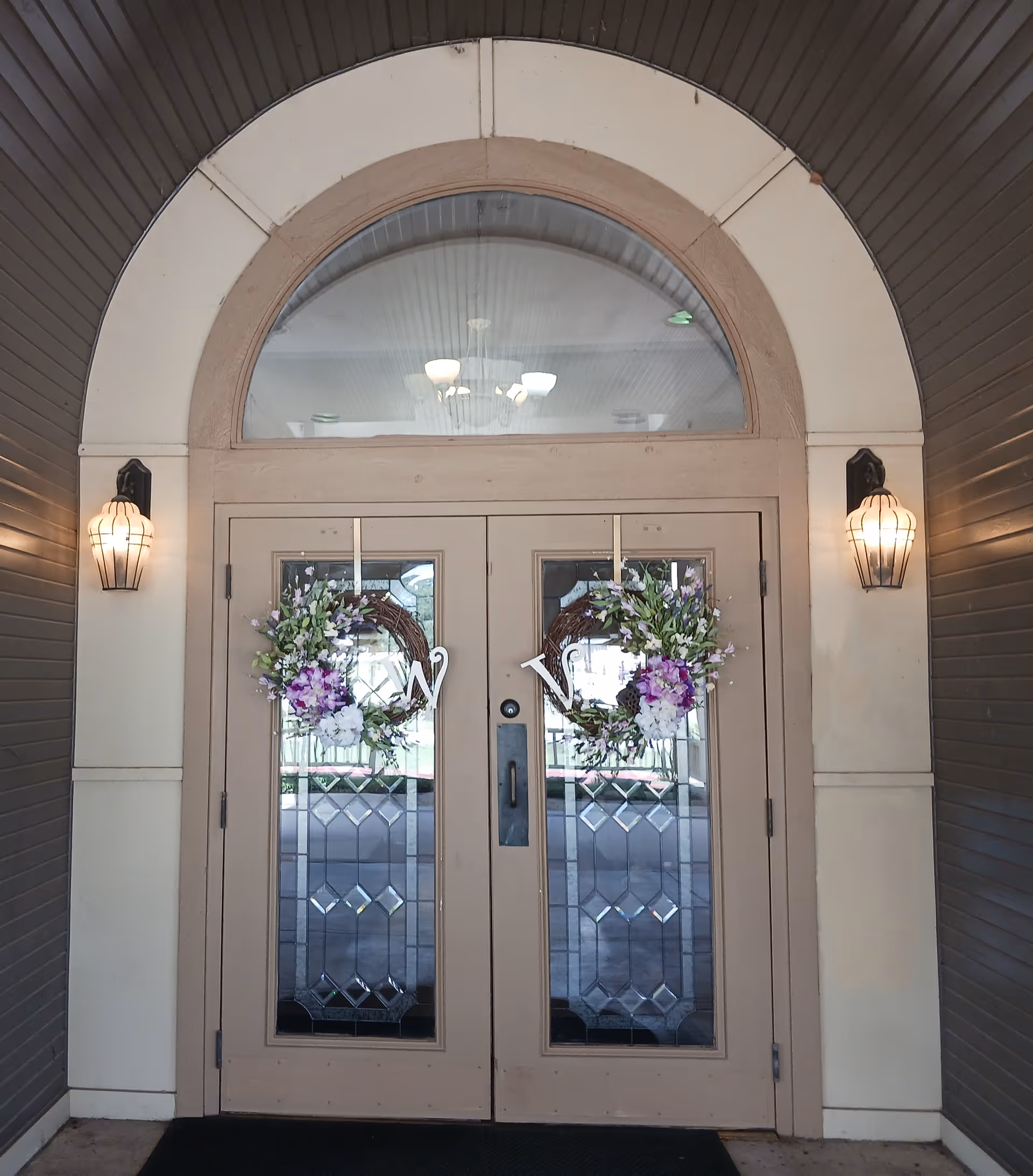 Double glass doors with decorative wreaths featuring purple and white flowers and a letter 'W' on each wreath. The doors are set in an arched entryway with two wall-mounted lantern-style lights on either side.