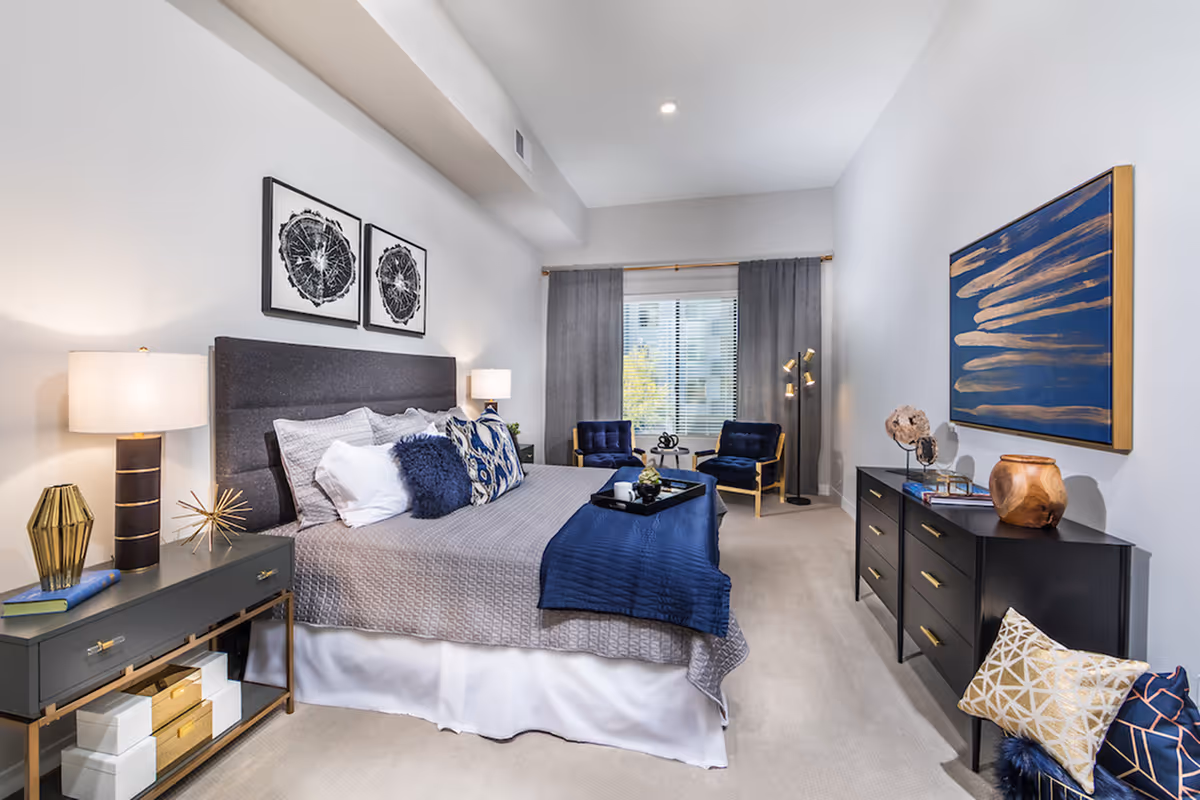 A modern bedroom with a large bed featuring gray and blue bedding, two bedside tables with lamps, two blue armchairs near a window with gray curtains, a black dresser with decorative items, and abstract artwork on the walls.