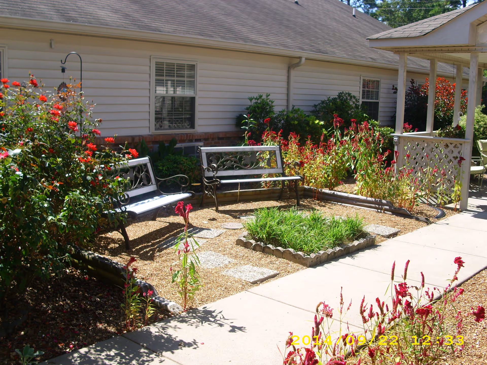Outdoor garden area at Magnolia Place Assisted Living featuring two metal benches with decorative backs, surrounded by various flowering plants and shrubs. A paved walkway runs alongside the garden, and the exterior wall of the building with windows is visible in the background.
