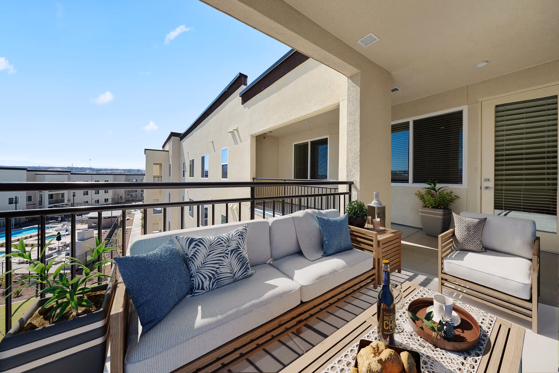 A covered outdoor balcony area with a cushioned sofa and armchair, decorated with blue and patterned throw pillows. A wooden coffee table holds a bottle, a cup, a tray with greenery, and a basket of bread rolls. Potted plants are placed around the seating area. The balcony overlooks a courtyard with a swimming pool and other buildings under a clear blue sky.