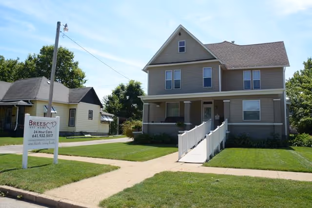 Front view of a two-story beige residential building with a wheelchair ramp, lawn, sidewalk and a yard sign for Brees Rest Home.