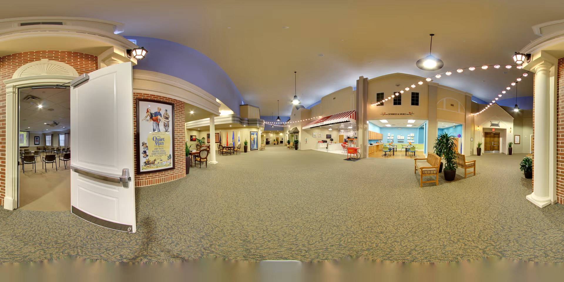 Wide panoramic view of a spacious indoor common area in a senior living facility. The area features carpeted floors, high ceilings with hanging lights and string lights, and a variety of seating options including chairs and benches. There are small storefront-style sections along the walls, including a market area and a room with tables and chairs. An open door on the left leads to a room set up with rows of chairs, possibly for meetings or events. The space is well-lit and decorated with plants and framed posters.