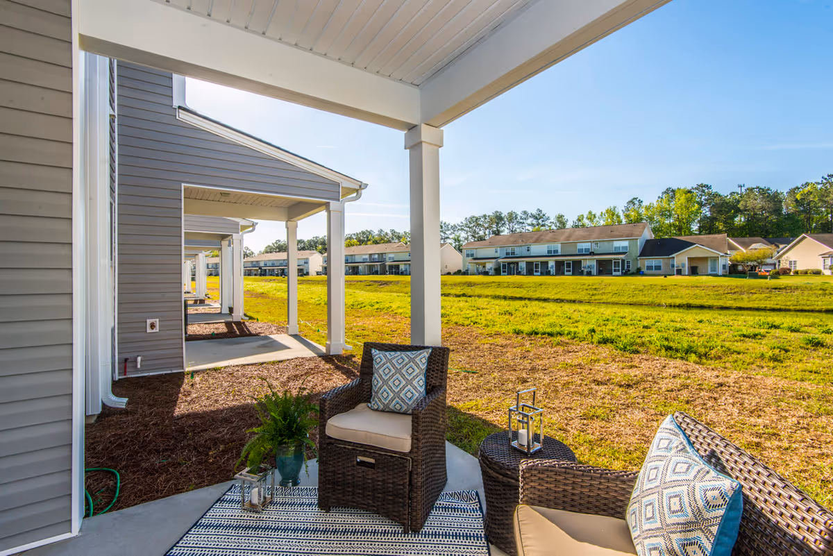 Covered outdoor patio area with two wicker chairs with cushions and a small round table with a lantern, overlooking a grassy open space and neighboring residential buildings under a clear blue sky.