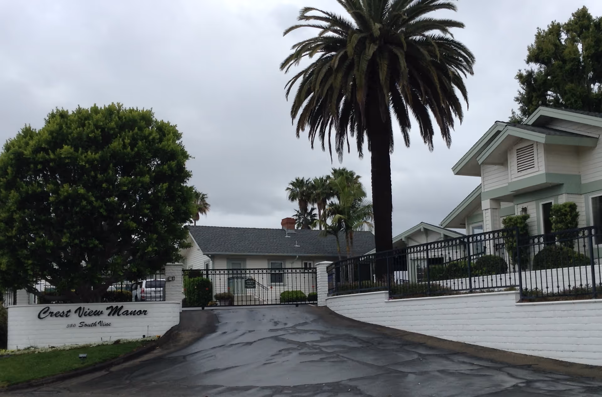 Entrance driveway to Crest View Manor, a residential facility with white buildings, a gated entrance, and lush landscaping including a large palm tree and other greenery under a cloudy sky.