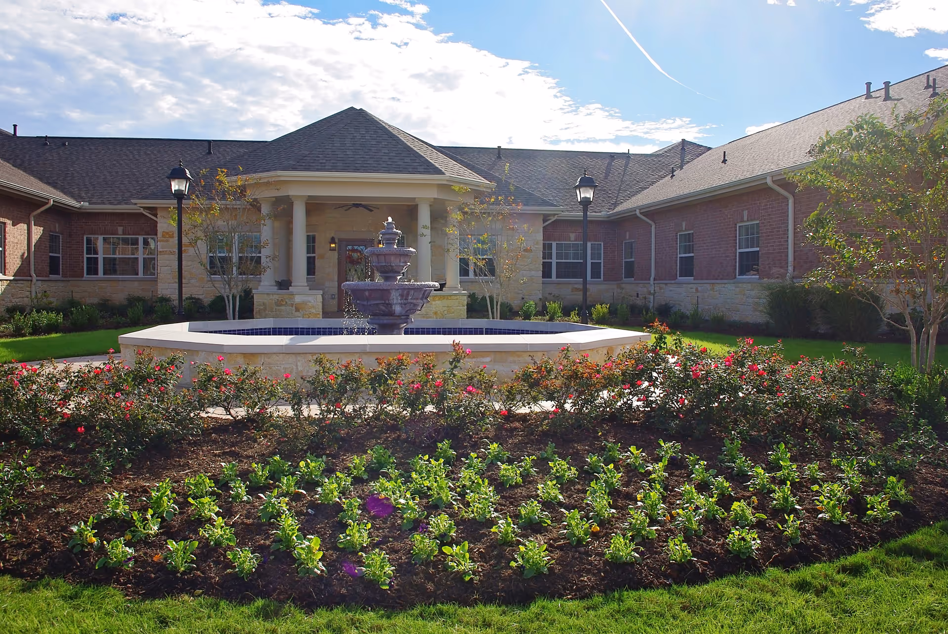 Outdoor view of Cypress Assisted Living - CyFair featuring a landscaped garden with blooming flowers and greenery in the foreground, a large decorative water fountain in the center, and a brick building with a covered entrance and columns in the background under a partly cloudy sky.