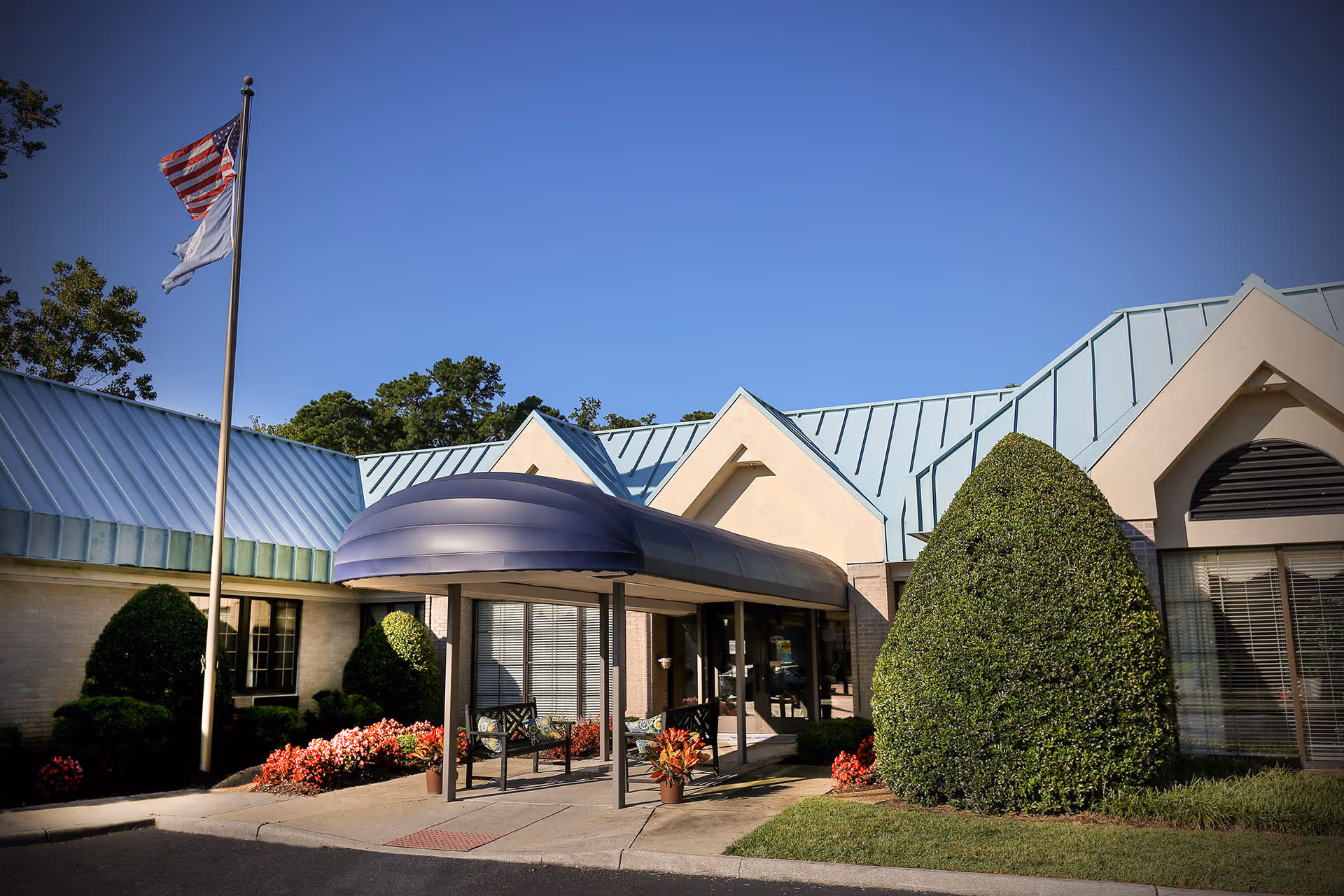 Exterior view of The Newport Nursing and Rehabilitation Center building entrance with a blue metal roof, a covered walkway, neatly trimmed bushes, colorful flower beds, and two flagpoles displaying the American flag and another flag against a clear blue sky.
