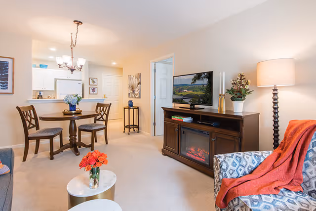 A cozy living room and dining area in a retirement community apartment. The living room features a patterned armchair with an orange throw, a wooden TV stand with an electric fireplace, a flat-screen TV, a floor lamp, and decorative plants. The dining area has a round wooden table with two chairs and a vase of flowers. The kitchen with white cabinets and appliances is visible in the background.