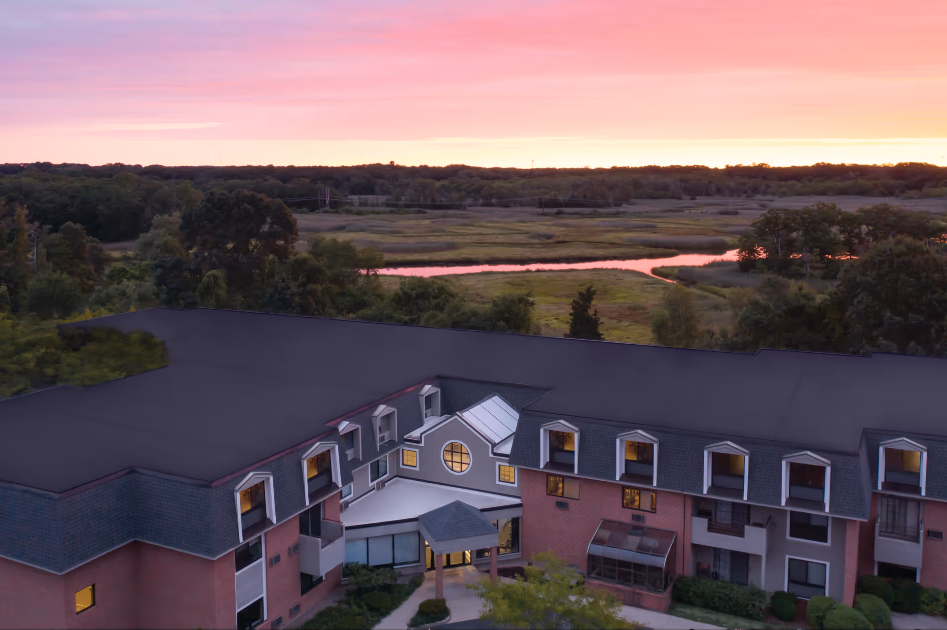 Aerial view of a senior living facility building with a dark roof and brick walls, surrounded by trees and greenery, with a river and forested area in the background under a pink and orange sunset sky.