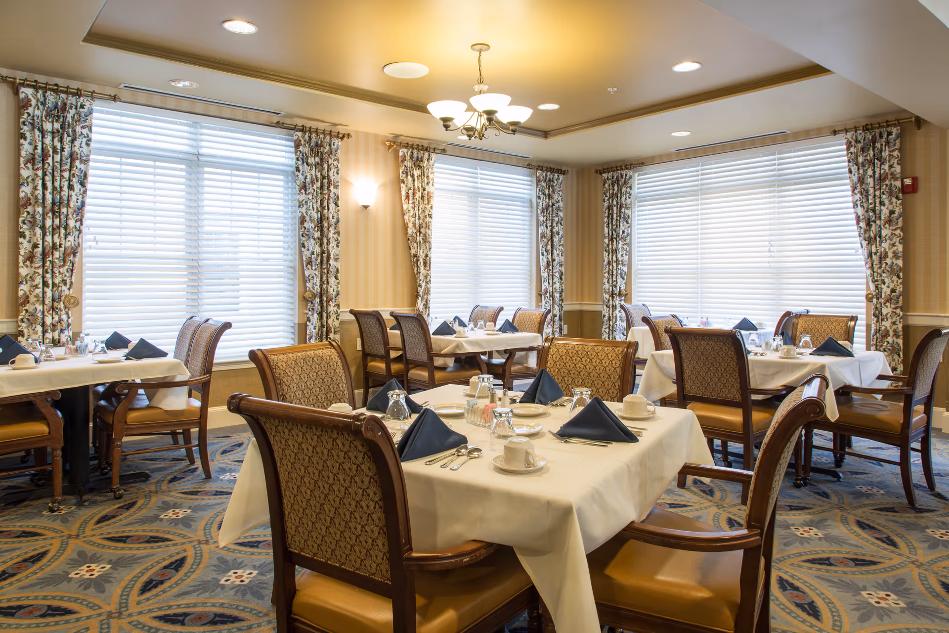 A dining room with several tables covered in white tablecloths, each set with cups, glasses, silverware, and navy blue folded napkins. The room has large windows with floral curtains and patterned carpet flooring. The lighting includes a chandelier and wall sconces.
