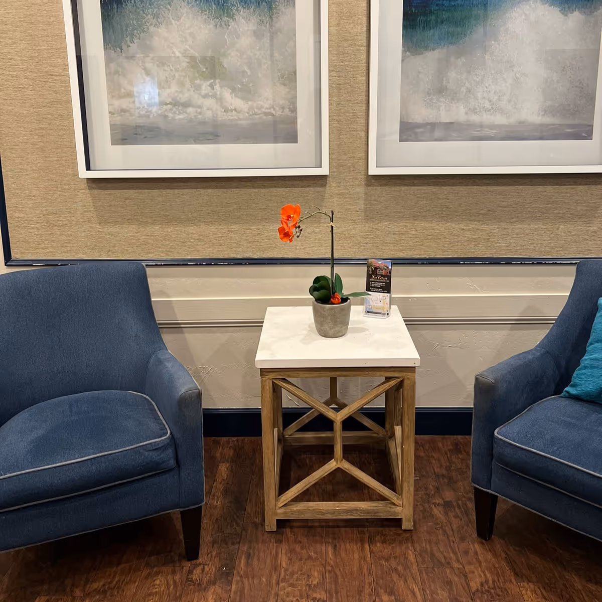 Two blue upholstered armchairs with a small wooden side table between them. The table has a white top and holds a small potted plant with orange flowers and a brochure. Behind the chairs are two framed pictures of ocean waves on a beige textured wall.