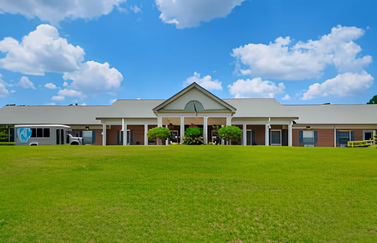 Front exterior view of a single-story building with a large green lawn in front, blue sky with scattered clouds above, and a bus parked on the left side.