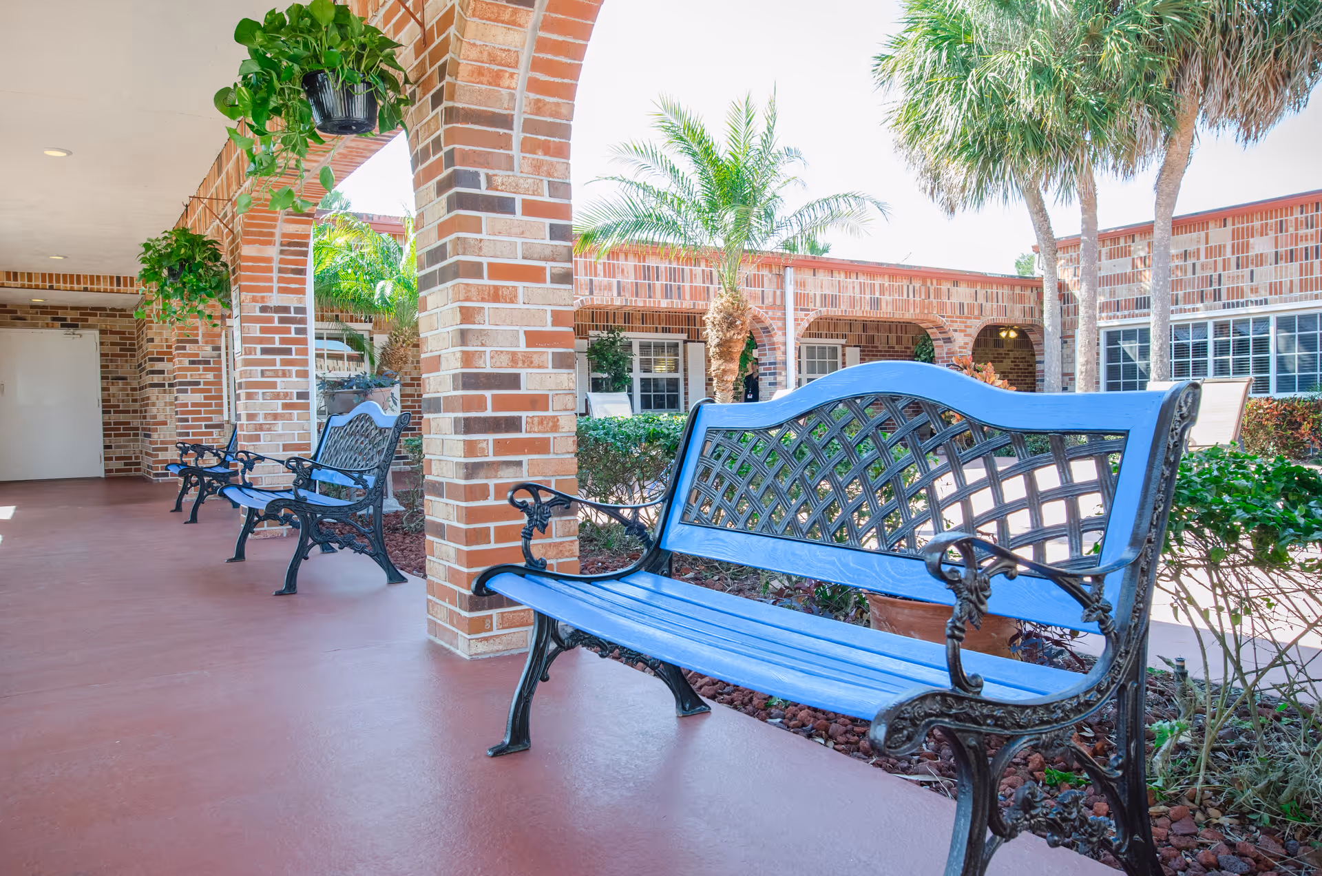Outdoor seating area at Anchor Care and Rehabilitation Center featuring blue metal benches with decorative backs, brick columns and arches, hanging potted plants, palm trees, and shrubbery under a covered walkway.