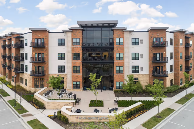 Front exterior of a modern four-story Rose Senior Living building with a central glass atrium, balconies, and a landscaped courtyard with outdoor seating.