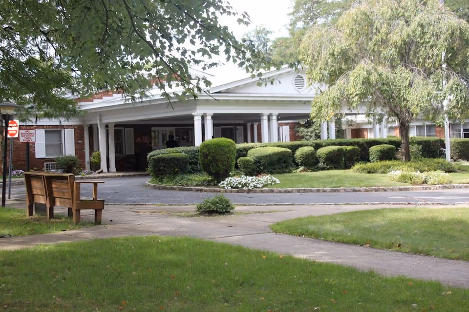 Front exterior view of Cornell Hall Care & Rehabilitation Center showing a single-story building with white columns, trimmed bushes, a tree, a bench, and a paved walkway surrounded by green grass.