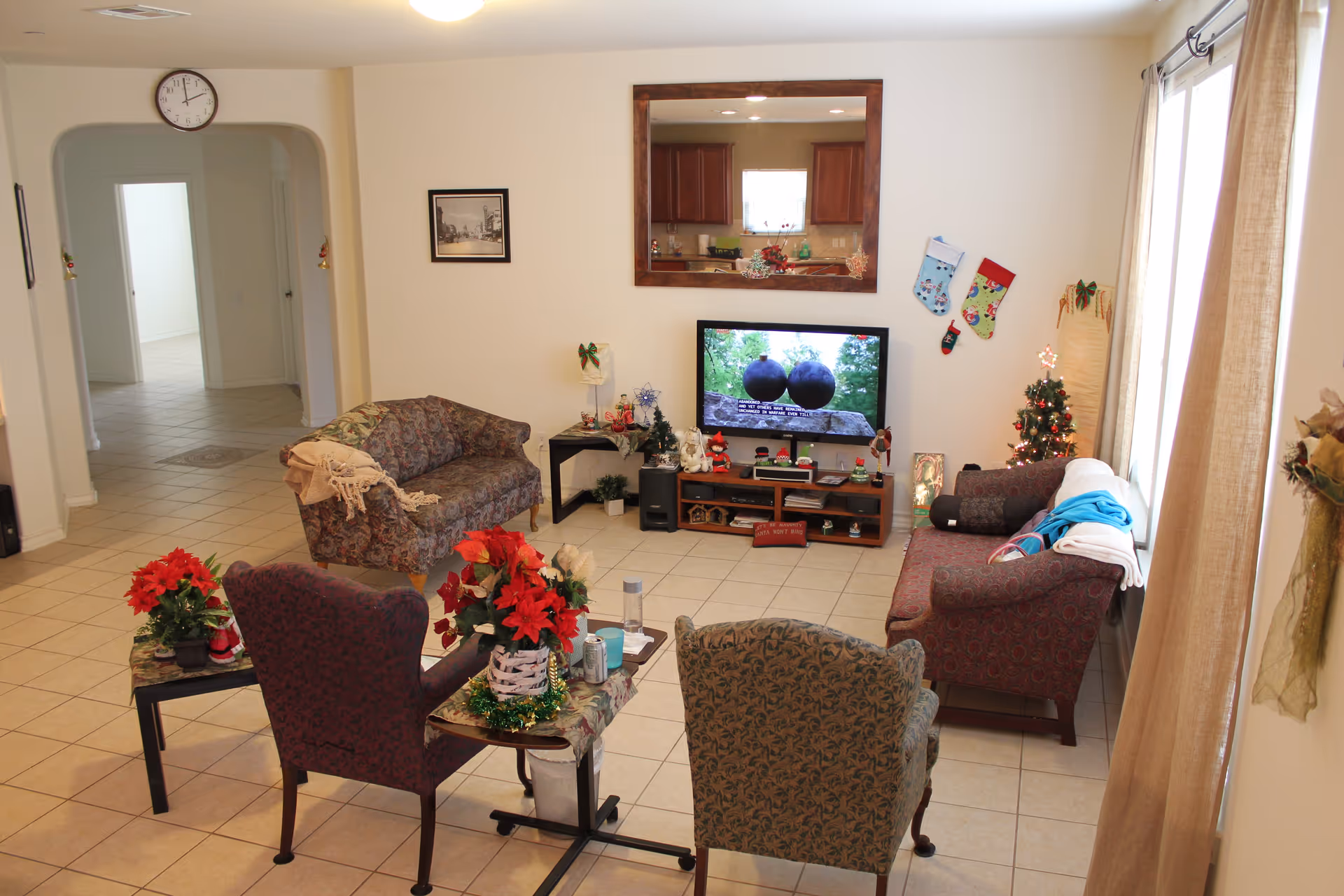 A cozy living room decorated for Christmas with a TV showing a nature scene, a small Christmas tree, stockings hanging on the wall, and four upholstered chairs arranged around a small table with poinsettia plants. There is a large mirror above the TV and a clock on the wall near an arched doorway leading to other rooms.
