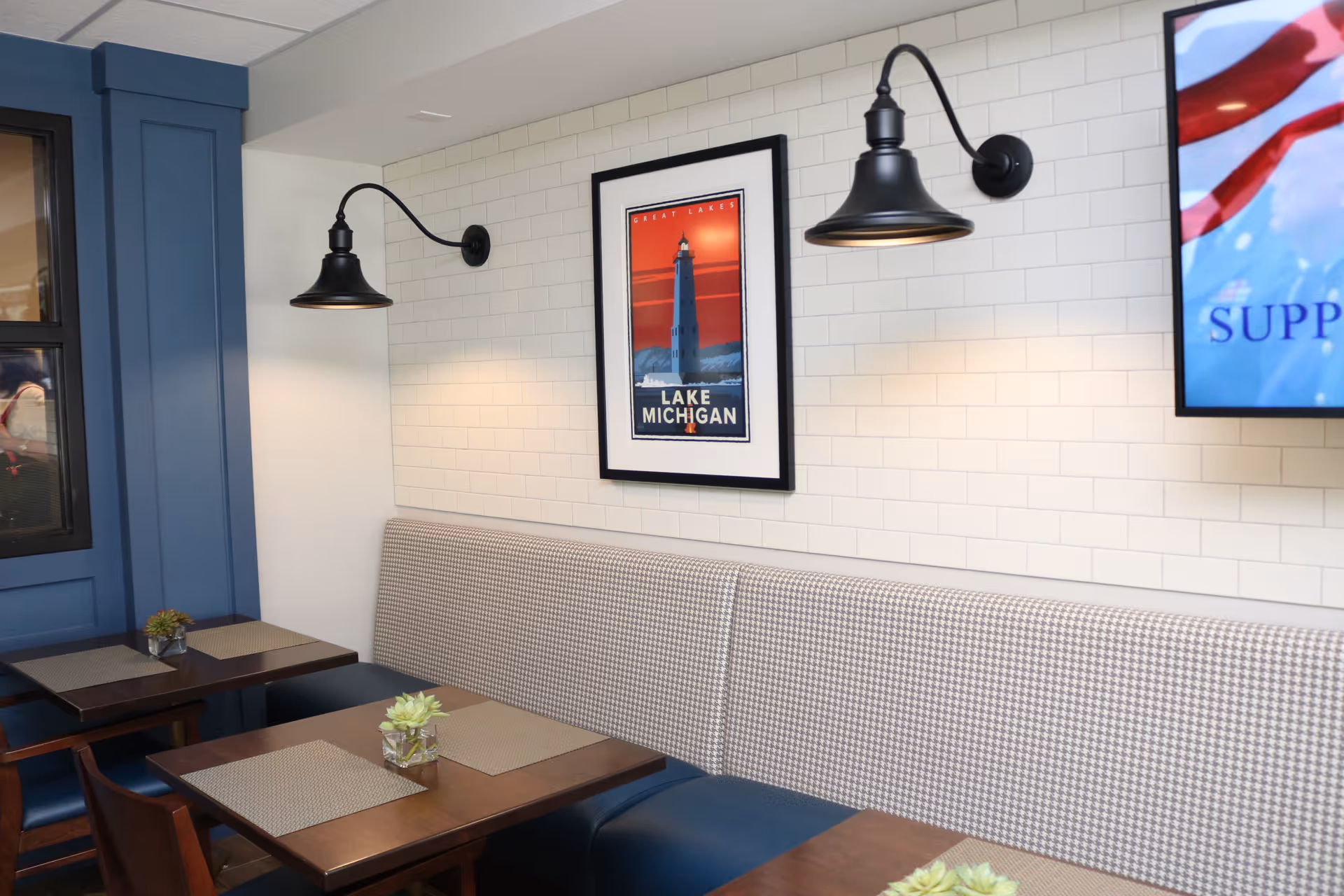 Interior view of a dining area with wooden tables, cushioned bench seating along a white tiled wall, two black wall-mounted lamps, a framed picture of a lighthouse with the text 'Lake Michigan', and a TV screen partially visible on the right.
