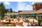 Outdoor patio area at a senior living facility with multiple tables and chairs, some shaded by green umbrellas, surrounded by buildings and trees under a blue sky with scattered clouds.