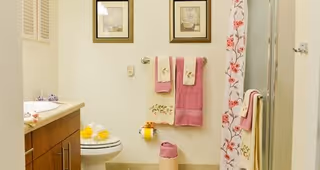 A bathroom with a wooden vanity and sink on the left, a toilet with yellow decorative items on top, and a shower with a floral-patterned curtain on the right. Two framed pictures hang on the wall above a towel rack holding pink and cream towels. A pink and cream wastebasket is on the floor.
