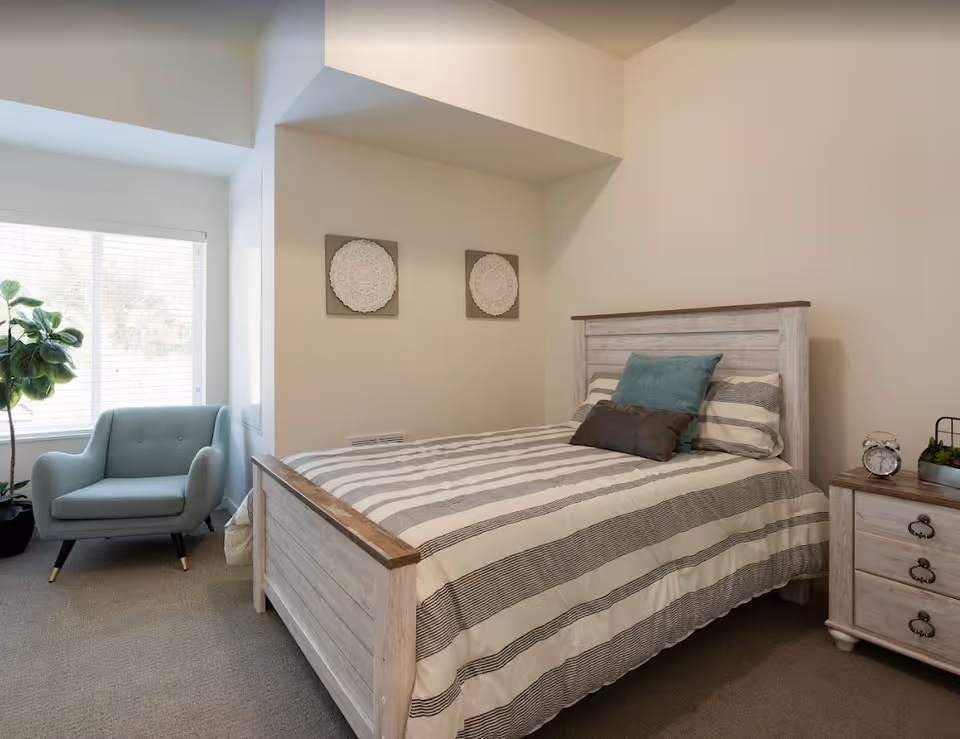 A cozy bedroom with a wooden bed frame and striped bedding, two decorative wall hangings above the bed, a light blue armchair near a window with blinds, a potted plant beside the chair, and a wooden nightstand with an alarm clock and a small plant.