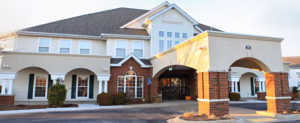 Exterior front view of a senior living facility building with a covered entrance supported by brick and white columns, multiple windows, and a sloped roof under a clear sky.