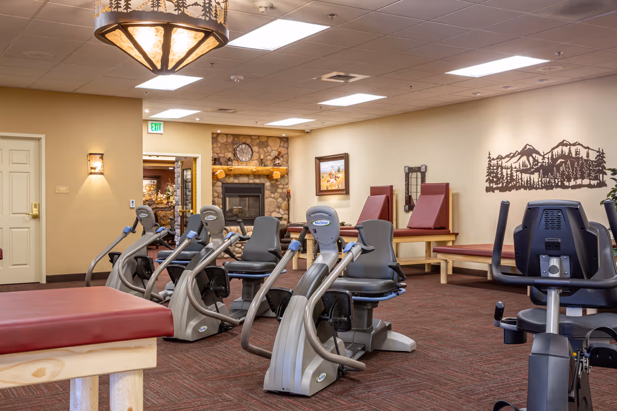 A well-lit exercise room in a senior living facility featuring several NuStep recumbent cross trainers arranged on a carpeted floor. The room has beige walls with a mountain and forest mural, a stone fireplace with a clock above it, and padded benches along the walls. A decorative ceiling light fixture and wall sconces provide additional lighting.