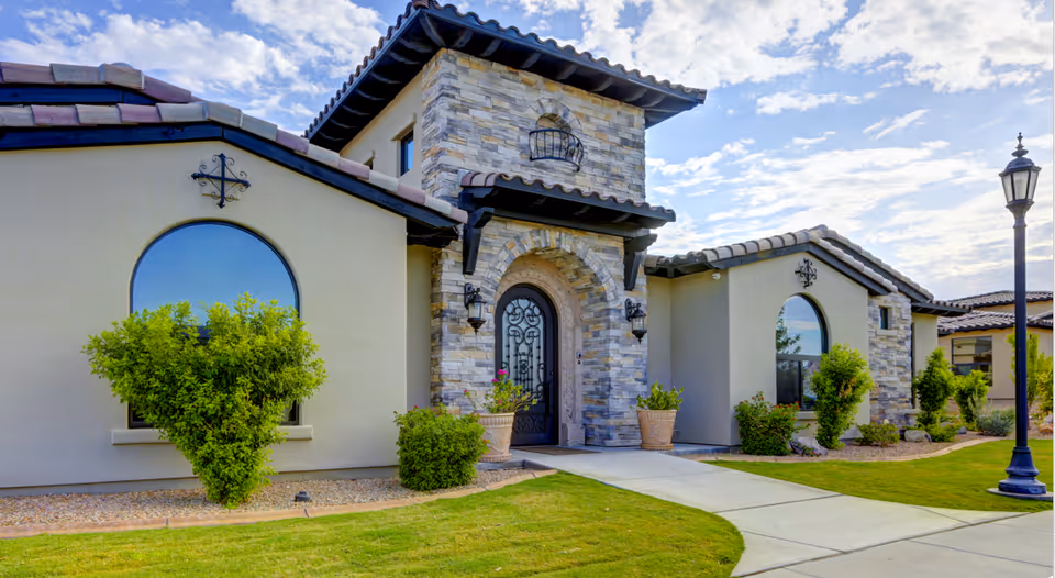 Mediterranean-style building front with a stone arched entrance, tiled roof, arched windows, landscaping and a paved walkway.