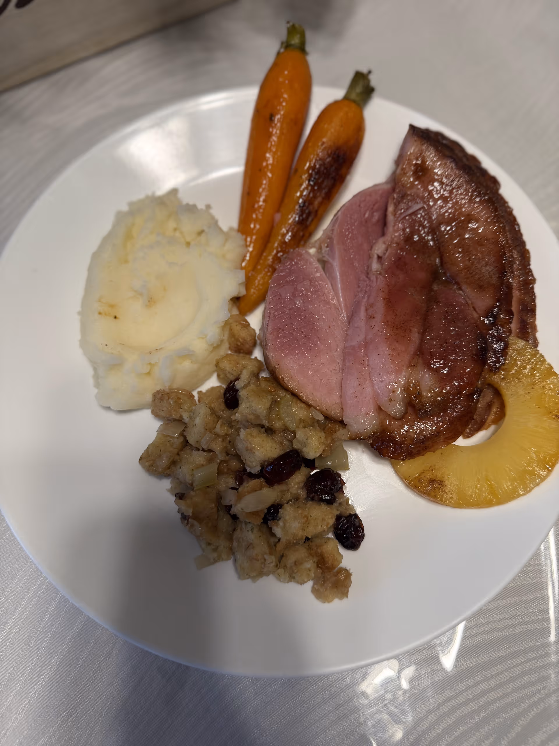 White plate with sliced ham, mashed potatoes, roasted carrots, stuffing with raisins, and a pineapple ring.