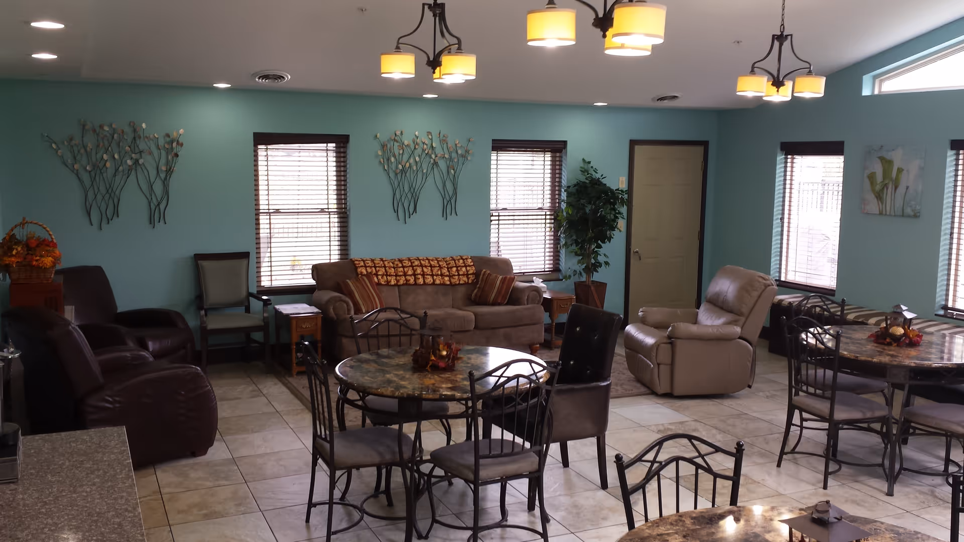 A senior living facility common area with teal walls, multiple windows with blinds, and tiled floors. The room contains several round tables with metal chairs, a brown sofa with patterned pillows, two recliner chairs, a green armchair, side tables, wall decorations, and ceiling light fixtures.