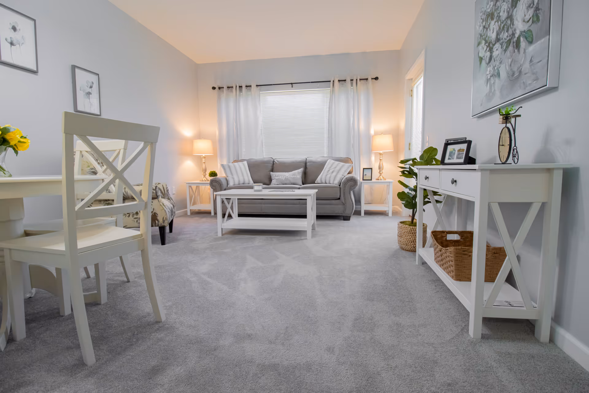 Airy living room with a gray sofa and pillows, white coffee and side tables with lamps, a dining chair in the foreground, and light gray carpet.