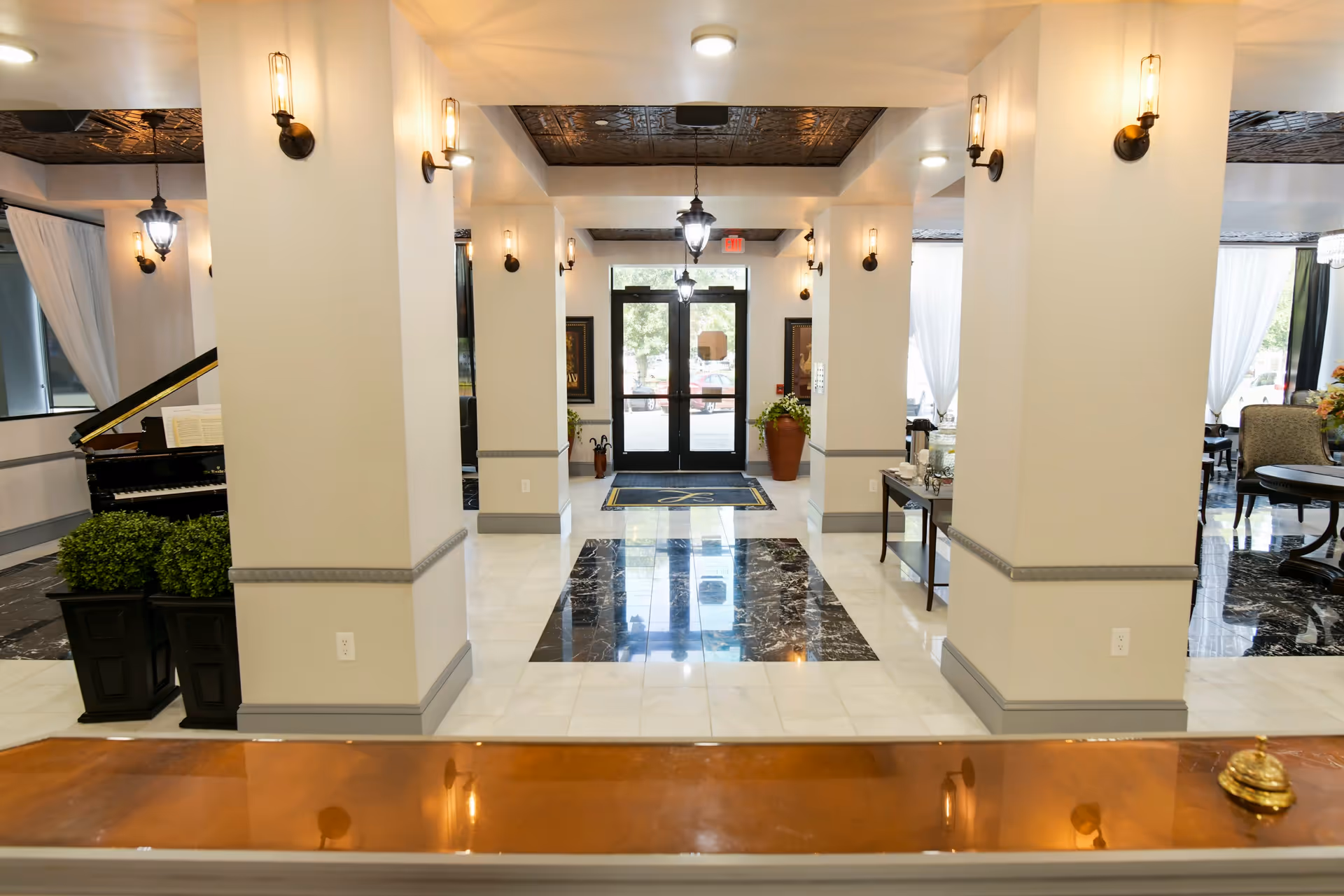 View of a bright and elegant assisted living facility lobby with white walls, black and white tiled floor, multiple columns, wall sconces, a grand piano on the left, and a seating area with chairs and tables on the right. The entrance door is visible at the far end with glass panels letting in natural light.