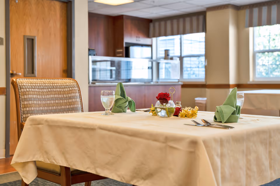 A dining table set with a beige tablecloth, two green folded napkins, two glasses of water, silverware, and a small vase with a red rose and yellow flowers in a well-lit room with windows and wooden furniture.