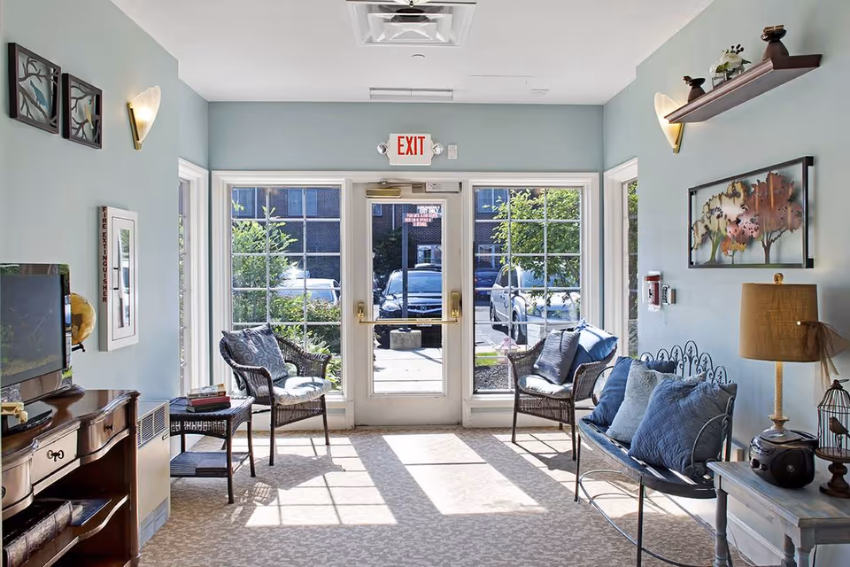 A bright and cozy sitting area near the entrance of a facility with large glass doors and windows letting in natural light. The room features two wicker chairs with cushions, a metal bench with blue pillows, a wooden side table with a lamp, and decorative wall art. Outside, cars and greenery are visible through the windows.