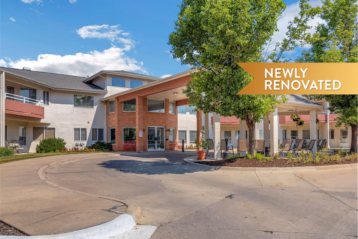 Front entrance of a senior living building with a covered portico, circular driveway, trees and outdoor seating.