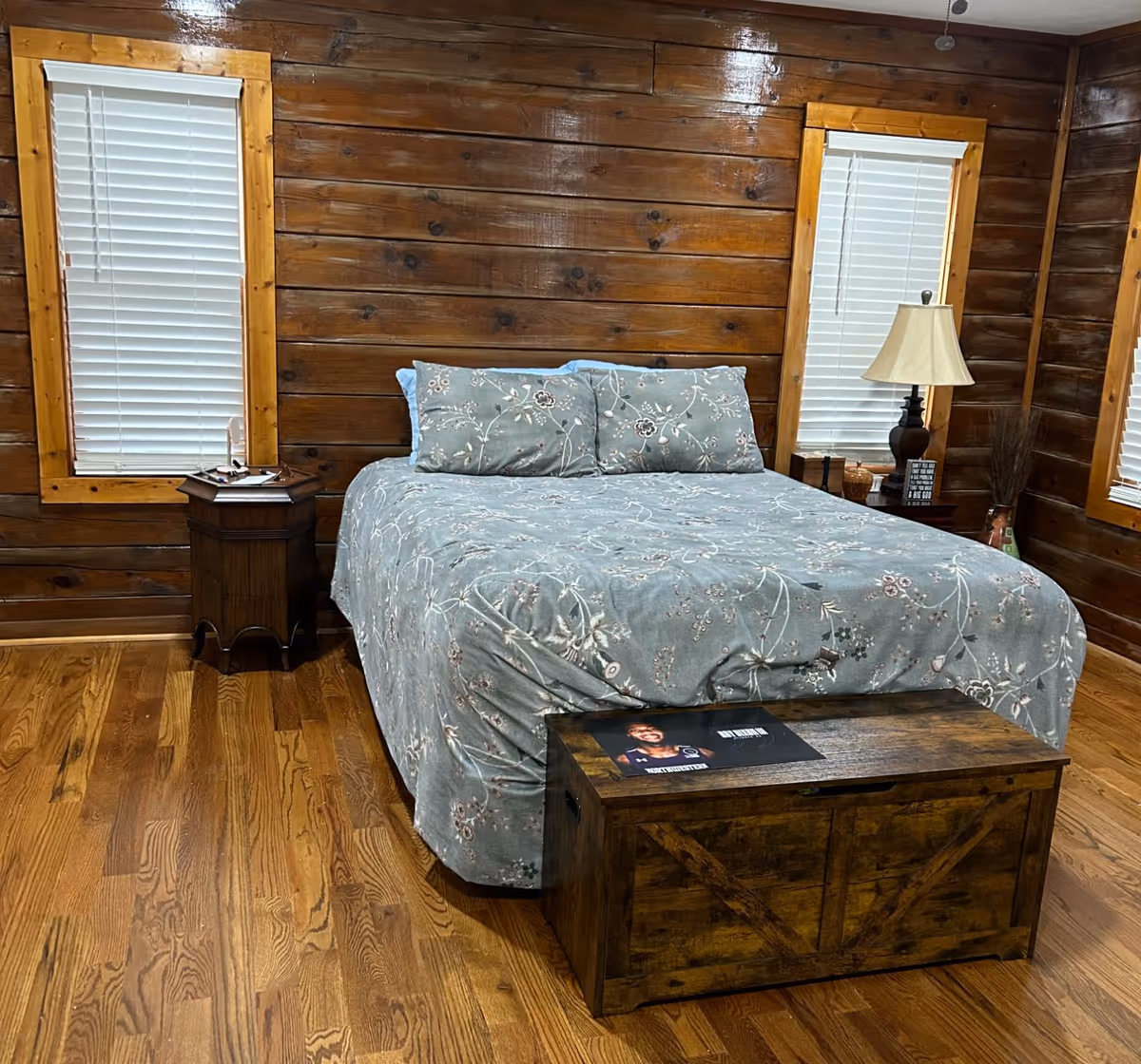 Cozy wooden bedroom with a bed dressed in a gray-blue floral comforter, two windows with blinds, bedside tables and a wooden chest at the foot.