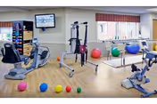A well-lit physical therapy or exercise room with various fitness equipment including stationary bikes, parallel bars for walking support, and colorful exercise balls scattered on a wooden floor. There is a wall-mounted TV and large windows with red valances allowing natural light into the room.
