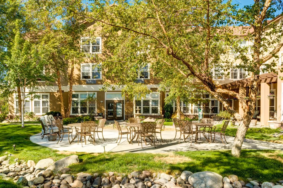 Outdoor patio area with metal tables and chairs arranged on a circular concrete surface surrounded by green grass, trees, and rocks in front of a multi-story building with many windows.