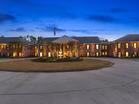 Exterior view of a large brick senior living facility building at dusk with lights on inside. The building has a central entrance with white columns and an American flag flying above. There is a circular driveway with a landscaped island in the center featuring trees and a fountain.