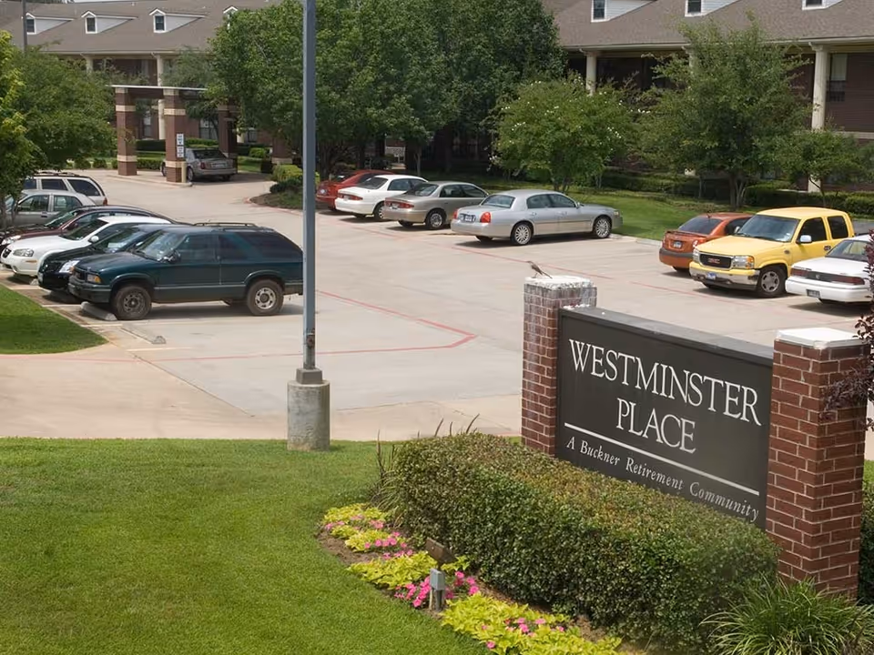 Parking lot with several parked cars in front of a building with a covered entrance. A sign in the foreground reads 'WESTMINSTER PLACE A Buckner Retirement Community' with landscaping including bushes and flowers around it.
