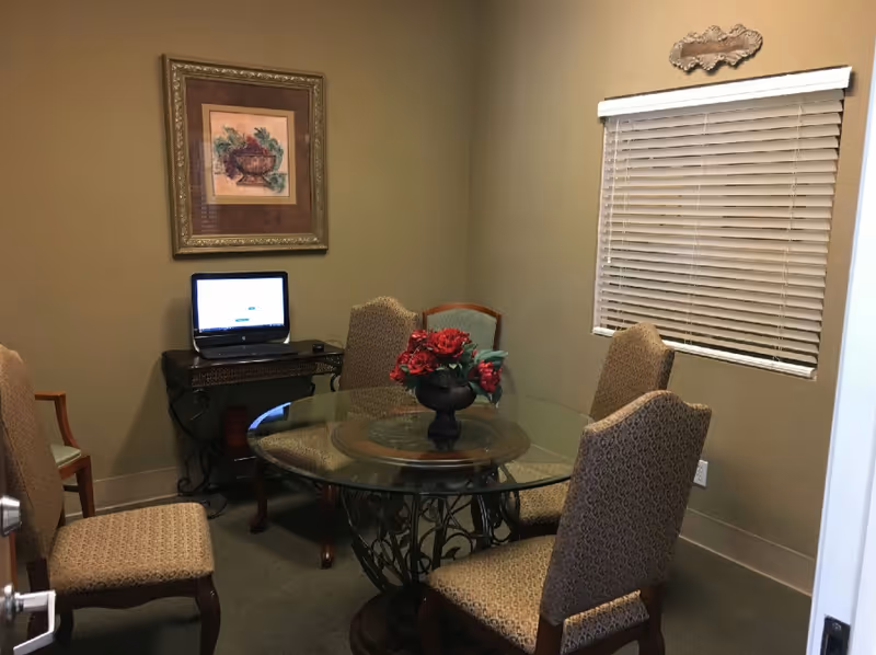 A small meeting or dining room with a round glass table surrounded by five upholstered chairs. A vase with red flowers is centered on the table. There is a window with closed blinds on the right wall and a framed painting of a plant on the left wall. A small table against the left wall holds an open laptop.